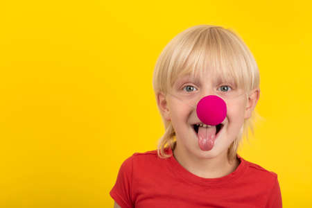 Blond boy with clown nose looking at camera and showing tongue. Portrait on yellow background.の写真素材