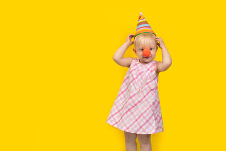 Little girl in party hat and red clown nose on yellow background. Childrens party.の写真素材