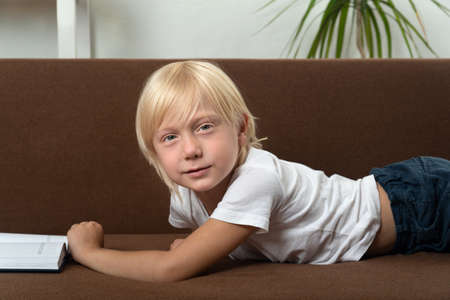 Blond schoolboy is lying on couch with book. Portrait of boy with textbook.の写真素材
