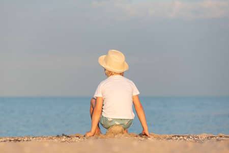 Boy in straw hat sitting on sandy beach and looks out to sea. Back view.の写真素材