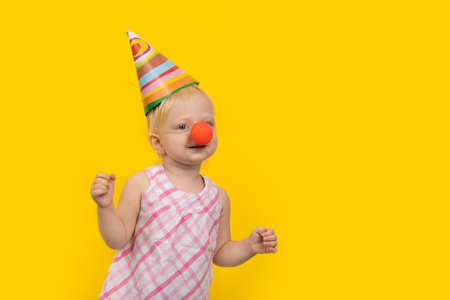 Little girl in festive hat and clown nose on yellow background