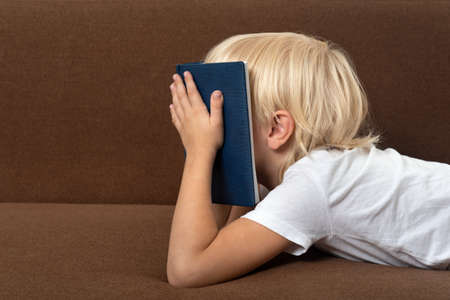 Boy lies on couch with book to his face. Child is tired of studying. Distance learning problems