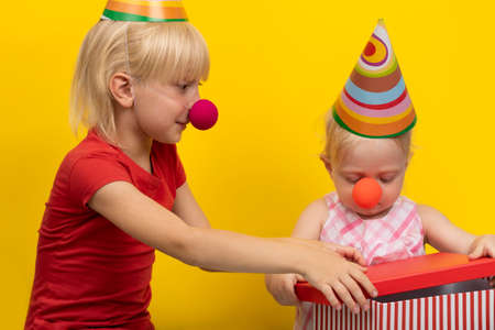 Children in festive hats and red clown noses in studio on yellow background. Children open gift.
