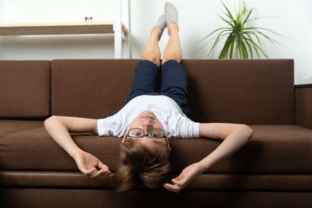 Child lies on couch with his head hanging down. Boy with glasses resting on sofa upside down.の写真素材