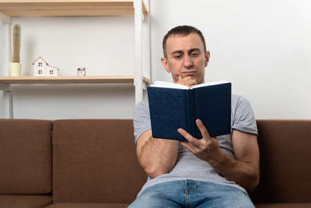 Young man thoughtfully reads book while sitting on sofa in living room