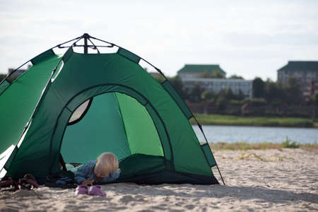 Small child lies in tourist tent against river and houses background.の写真素材