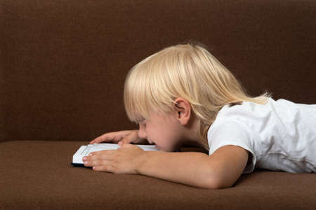 Blond boy lies on couch next to textbook. Boy is tired of studying. Homeschooling