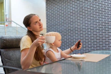 Mom with child in cafe. Mom thoughtfully drinks coffee with baby in her arms. Baby watching cartoon.の写真素材
