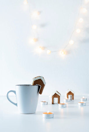 Cup of tea with gingerbread house and candles on white background. Christmas lights. Vertical frame.の写真素材