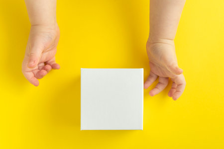 Children's hands and white cardboard box on yellow background top view. Copy space. Mock upの写真素材