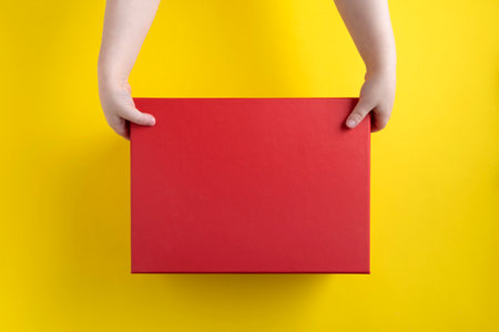 Children's hands opening red cardboard box. Yellow background. Top view. Copy space. Mock up.の写真素材