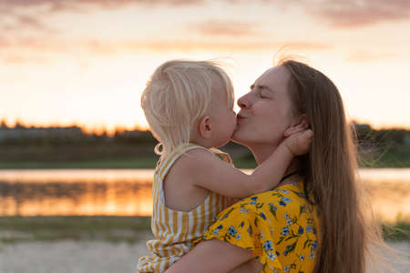 Young mother holding little daughter and gently kisses her. Portrait in sunset on river background.の写真素材