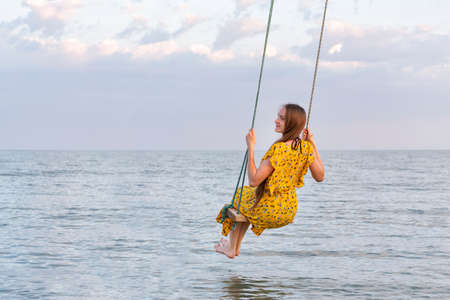 Beautiful woman in yellow dress sits on a rope swing over sea. Calm relax loneliness.の写真素材
