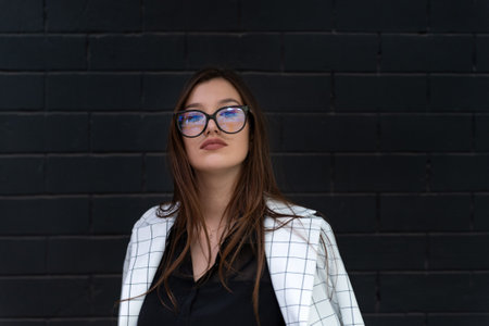 Portrait of stylish girl in glasses and jacket on black brick wall background.の写真素材