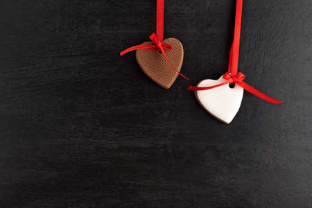 Two gingerbread heart-shaped cookies hang from red ribbon, black background. Mothers day. Womans...