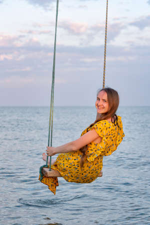 Young beautiful girl sitting on a rope swing on sea background. Vertical frameの写真素材