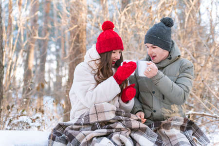 Love story. Young couple in winter park is wrapped in blanket and drinking teaの写真素材