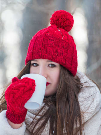 Beautiful brunette girl in red hat and mittens with mug of tea outdoors. Vertical frame portrait.