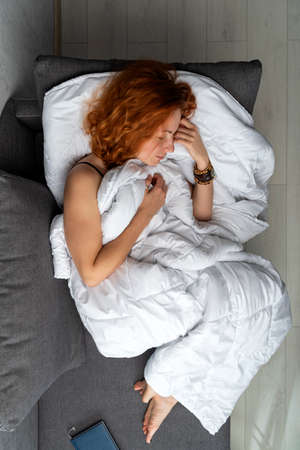 Beautiful young red-haired girl sleeps on the sofa, bypassing white blanket. Top view.の写真素材
