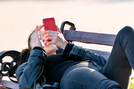 Girl lies on bench with phone in her hands. Young woman is resting in the park on the benchの写真素材