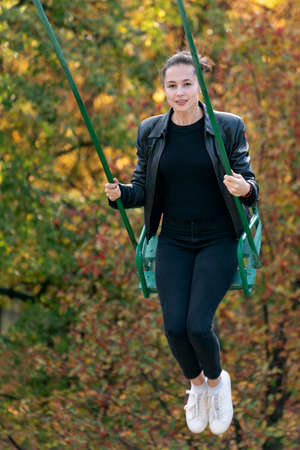 Young cheerful woman goes for a drive on swing in autumn park. Vertical frame.の写真素材