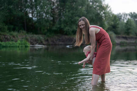 Mom and baby go into the river. Summer vacation with children. Child safety by the waterの写真素材