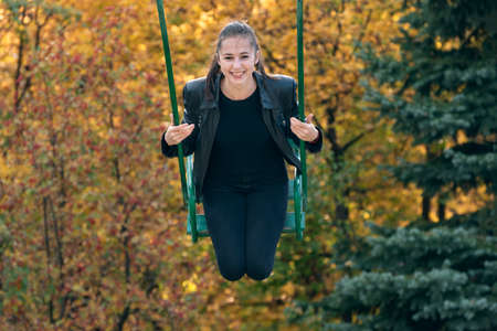 Young cheerful woman goes for a drive on swing on autumn park backgroundの写真素材