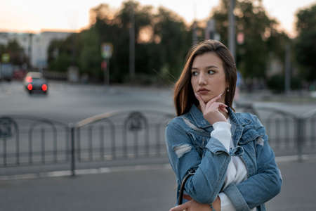 Young girl is waiting for someone by the road. Portrait of student on city background. Evening walk.の写真素材