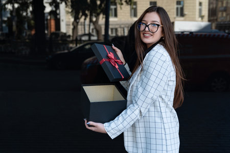 Brunette woman opened gift box looking at camera and smiling. Happy girl with gift on the street.