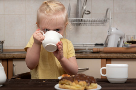 Child in the kitchen drinks from cup and looks at the cakes. Breakfast with child. Little girl drinking tea with pastry.の写真素材