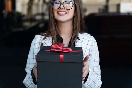 Young stylish woman hands gift. Happy young woman holds gift box and smiling. Black gift box with red bow.の写真素材