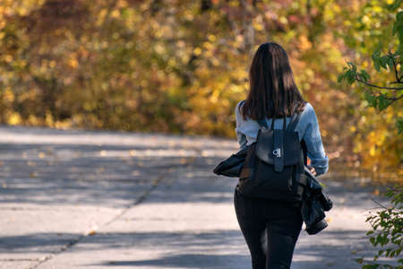 Brunette woman walks along the alley of an autumn park. Lonely girl in the sunny forest. Back viewの写真素材