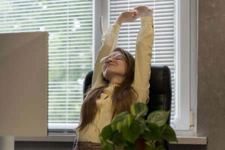 Young woman is stretching at workplace. Girl sits on chair and raised her arms up to stretch her...