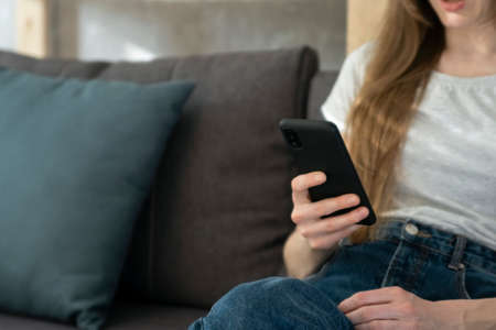 Girl is holding phone while sitting on sofa in living room. Close up. Woman is reading news or watching social networkの写真素材