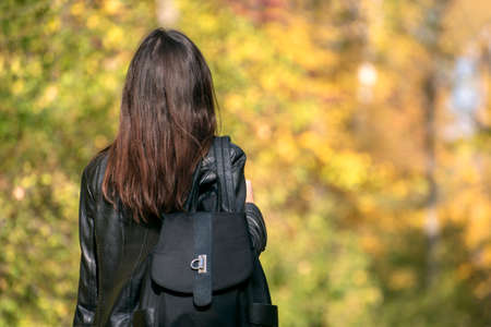Brunette girl with backpack walks in the park. Young woman in the forest. Back view.