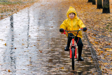 Gloomy boy in a yellow raincoat rides a bicycle along a wet alley in the rain. Autumn day.の写真素材