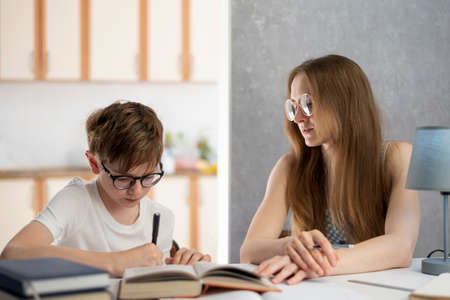 Boy with glasses is hard at work writing his homework. A young tutor helps the student with the lessons. Home schooling. Remote educationの写真素材