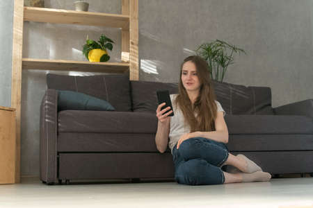 Girl looks with surprise at the phone screen. Young woman sitting at home with phone on floor and...