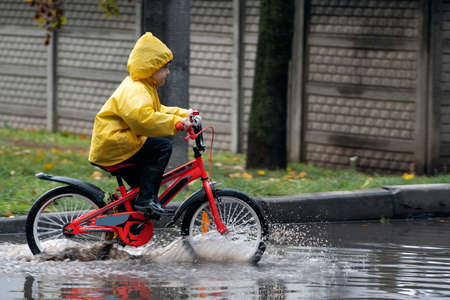 Happy boy in raincoat is fun rides on bicycle through the puddles. Child on bicycle in the rainの写真素材