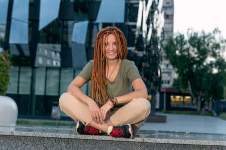 Woman with red dreadlocks in the lotus pose sits on a gray parapet and smiles. Against the background of an office building. Outdoorの写真素材