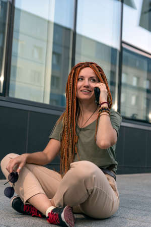 Beautiful girl with red dreadlocks talking on the phone and holding glasses in her hands. Against the background of an office building. Outdoors.の写真素材