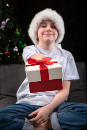 Boy in Santa hat is holding out present to you sitting on the sofa near the Christmas tree. New Year and Christmas. Vertical frame.の写真素材