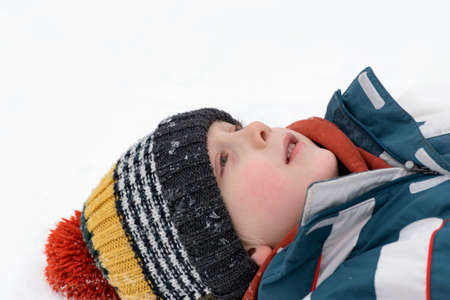 Portrait of boy lying in snow and looking up into sky. Child is wearing multi-colored knitted hat....