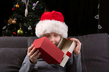 Child in Santa hat opens New Years gift sitting on the sofa against the background of the Christmas tree. Christmas eveの写真素材