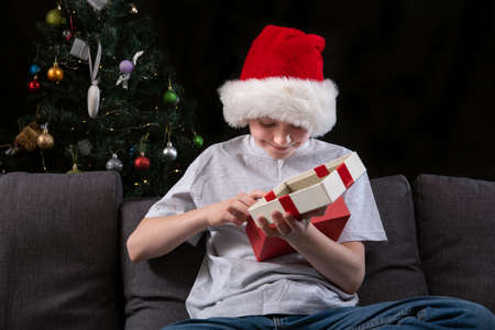 Happy boy in Santa hat opens gift box sitting on the sofa against the background of the Christmas tree. New Year giftsの写真素材