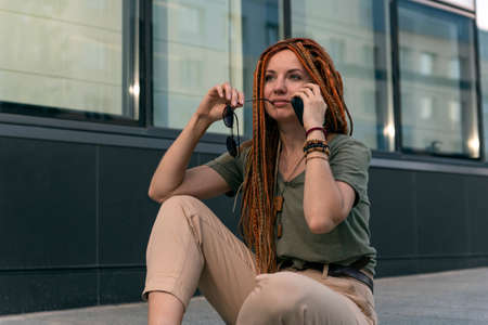 A young girl with red dreadlocks thoughtfully looks into the distance. Talking on the phone. Outdoors. High quality photoの写真素材
