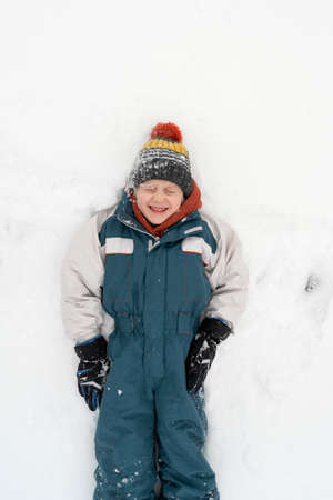 Grimacing boy in a warm jumpsuit and hat lies in the snow. Winter holidays. Child is playing in the snow. Top view.の写真素材