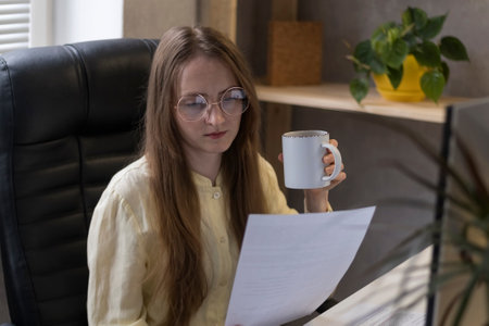 Young director in glasses is sitting in chair, drinking coffee and reading documents. Chief woman...