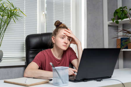 Young woman freelancer works from home. Girl sits at the workplace, works at laptop, tired of taking up her head.の写真素材