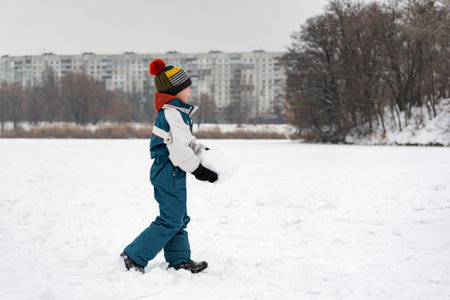 Child walks through snow-kept field. Boy in warm clothes plays outside in winter against the...